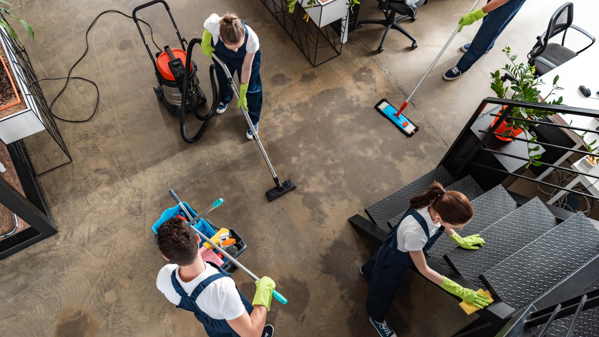 Aerial view of four people in uniform cleaning a modern office space. Two individuals are using mops and a vacuum, while another person is cleaning stairs and the fourth is managing cleaning equipment. The area features plants and contemporary furniture.