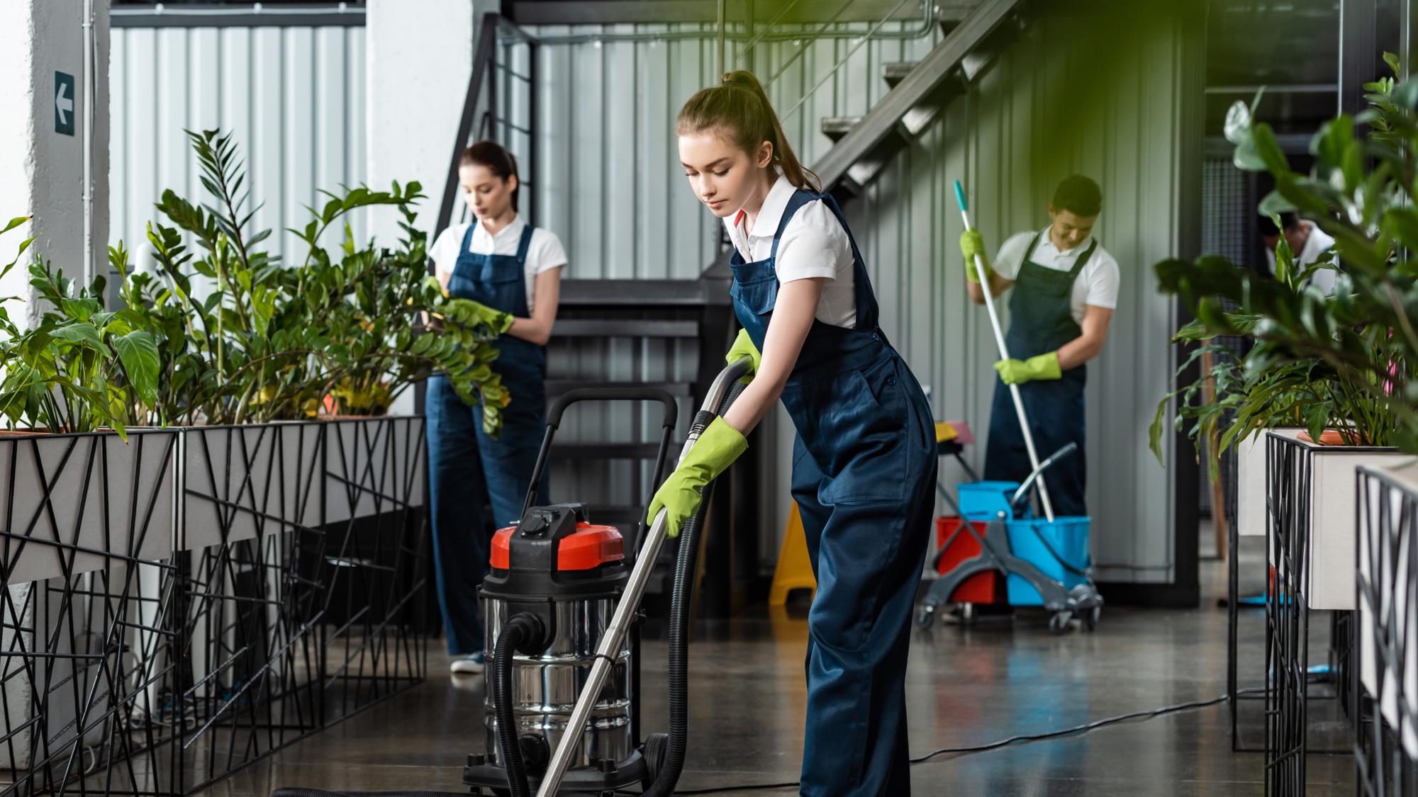 Two women in matching uniforms cleaning a modern indoor space with plants. One woman is using a vacuum while another in the background mops the floor. They are wearing gloves and the area is well-lit with a ladder visible in the background.